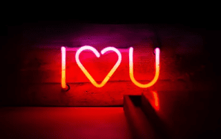 Neon sign displaying "i love you" with a heart symbol replacing the word "love," glowing in pink and red hues against a dark wooden background.