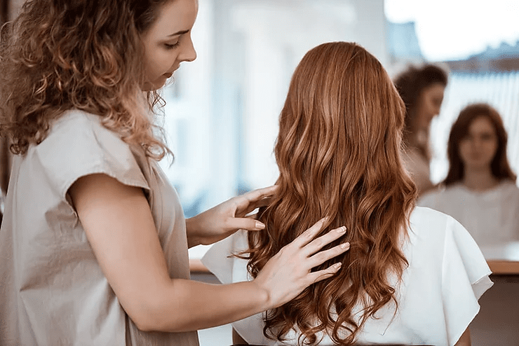 A hairstylist working on a woman's long curly hair in a salon, both reflected in a mirror in the background.