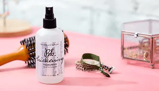 A bottle of hairspray labeled "oh. thickening" on a pink surface with a hairbrush and leopard print scrunchie in the background.