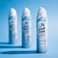Three cans of "bb scalp detox" foam lined up against a blue background, with the frontmost can displaying a swirl of white foam on its nozzle.