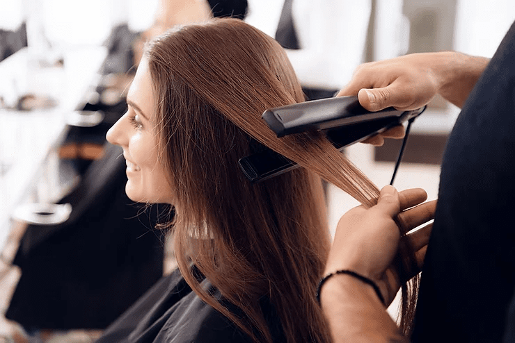A woman in a salon smiling as a stylist straightens her long brown hair with a flat iron. the setting shows other salon chairs blurred in the background.