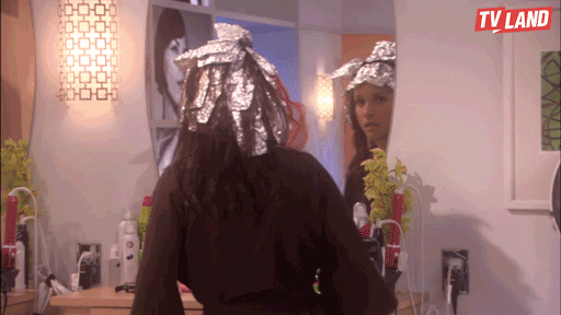 A woman in a kitchen is startled as she notices another person wearing an elaborate hat made of aluminum foil, who sneaks up behind her; both are reflected in a mirror.