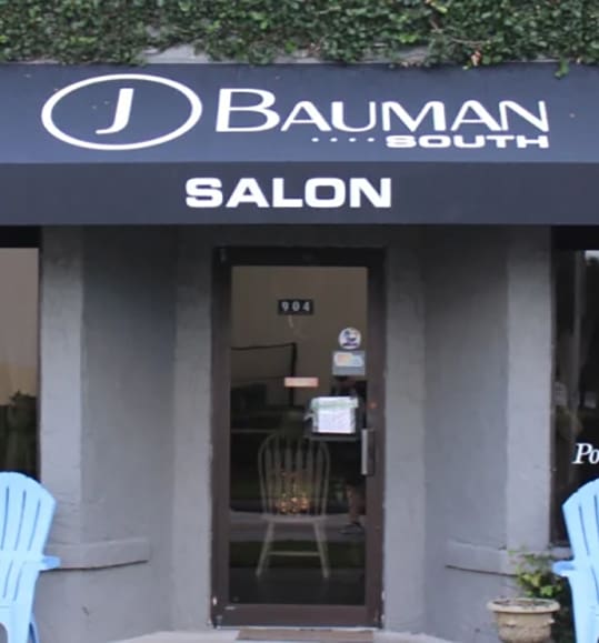 Entrance of the "j bauman salon south" with an open door, showing a glimpse of the interior. the entrance is flanked by light blue chairs, under a black awning with white text.