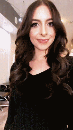 A woman with long, wavy brown hair, wearing a black top, smiles and poses in a salon.