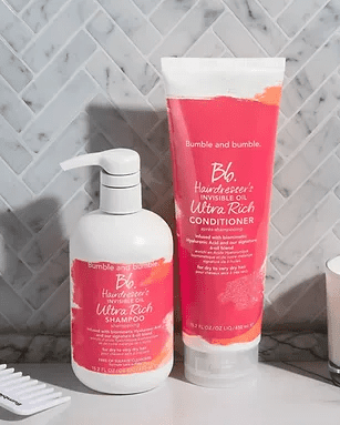 Two hair care products by bumble and bumble, a shampoo in a white pump bottle and a conditioner in a pink squeeze tube, are set against a gray tiled background with herringbone pattern.