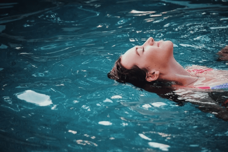 A woman relaxes by floating on her back in a serene blue pool, her eyes closed and face partially submerged in water, conveying a sense of peacefulness.
