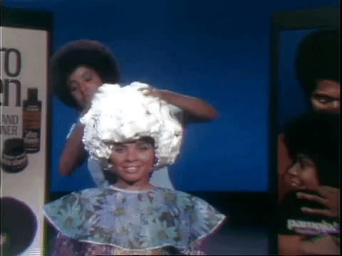 A vintage tv advertisement showing two people, one placing an oversized decorative wig on another's head, who is smiling and wearing a floral outfit. a product display is visible in the background.