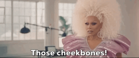 Rupaul, wearing a glamorous pink outfit and a voluminous white wig, gestures with admiration and says "those cheekbones!" in a gif.
