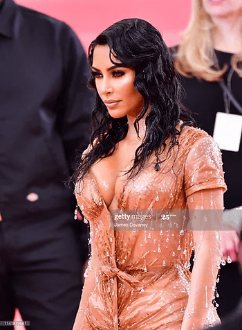 Kim kardashian wearing a shimmering, wet-look peach gown with deep neckline and beaded embellishments, posing at a red carpet event. her dark wavy hair complements her dramatic makeup.
