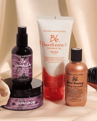 A collection of bumble and bumble hair care products displayed against a draped beige fabric background. items include sprays, a mask, and a treatment oil, all in stylish packaging.