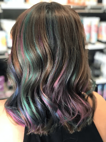 A woman's glossy, wavy hair with hidden rainbow highlights of blue, green, and purple expertly blended into her dark brown hair. the photo is taken from the back in a salon setting.