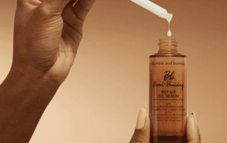 A person's hand holding a bottle of bumble and bumble bond-building repair oil serum with another hand dripping the serum from a dropper above it against a tan background.