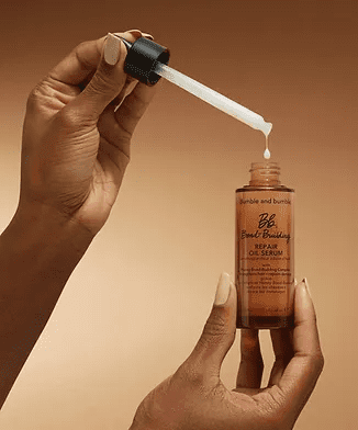 A hand holding a dropper dispenses a clear, viscous liquid into a small amber glass bottle labeled as "be smooth baking oil serum" against a neutral background.