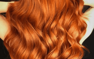 A close-up view of a person with vibrant, shiny copper-red hair styled in soft, flowing waves, highlighting its texture and color against a dark background.