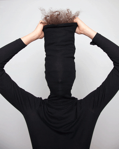 A person wearing a black turtleneck pulled over their head, with curly hair sticking out at the top, creating an amusing and whimsical appearance against a grey background.