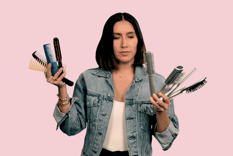A woman in a denim jacket juggles several hairbrushes with a confused expression against a pink background.