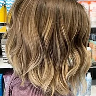 A close-up of a woman's hair styled in soft, voluminous waves, showcasing a blend of light and dark blonde highlights.