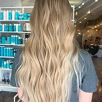 A woman with long blonde wavy hair sits in a hair salon, her back to the camera, showcasing her hairstyle against a backdrop of shelves filled with hair care products.