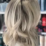 Blonde woman with shoulder-length, wavy hair highlighted with lighter blonde balayage, viewed from the back in a salon setting.