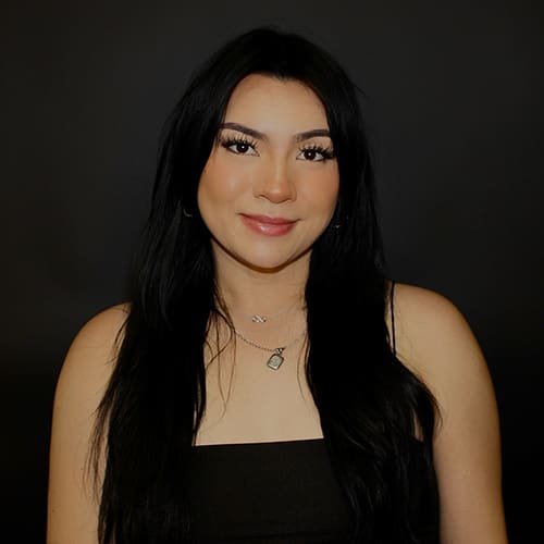 Portrait of a smiling woman with long straight black hair, wearing a black top and a necklace, against a dark background.