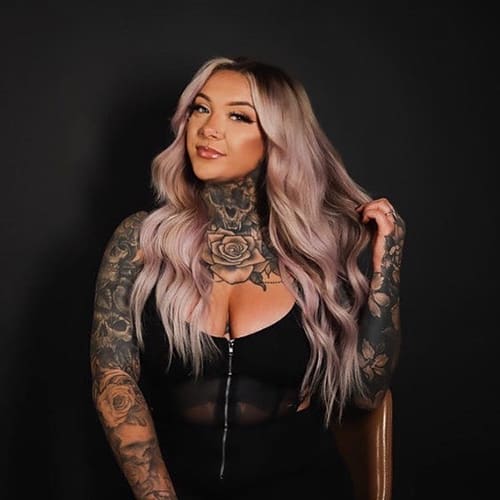A woman with long, wavy lavender hair and elaborate tattoos covering her arms and chest poses in a black top against a dark background.