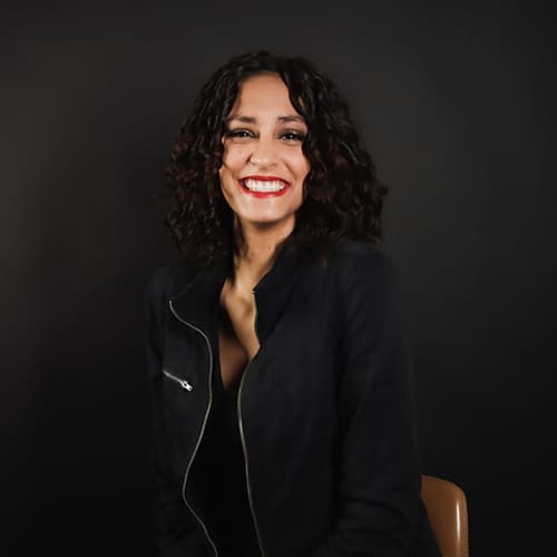 A woman with curly brown hair and red lipstick smiles warmly, wearing a black blazer. she is seated against a dark background.