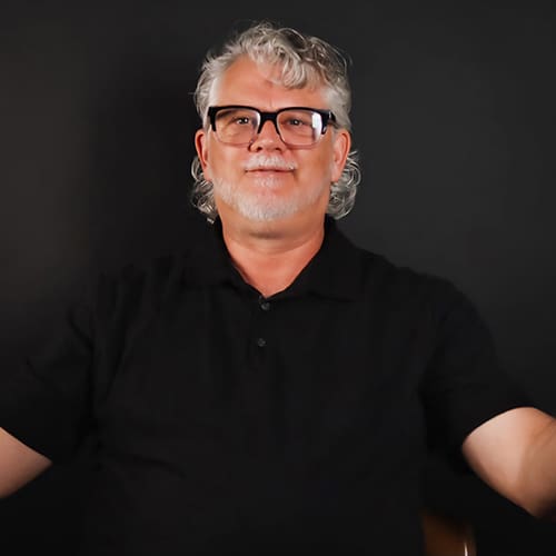 A middle-aged man with gray hair and glasses, smiling and wearing a black polo shirt, sitting against a dark background. his arms are spread slightly in a welcoming gesture.