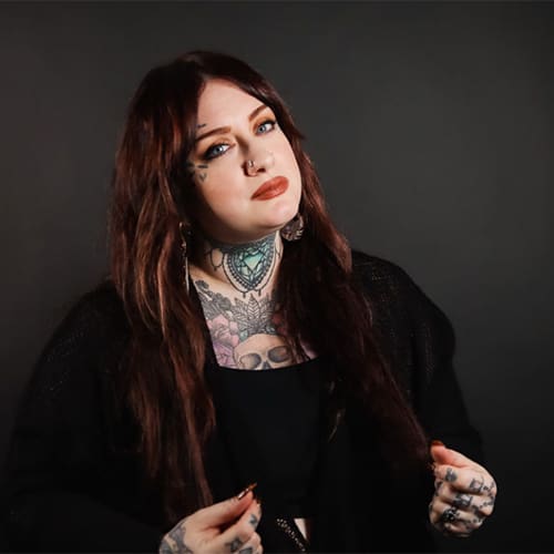 A portrait of a woman with long brown hair, facial and body tattoos, wearing a black outfit, posed against a dark background. she looks directly at the camera with a slight expression.