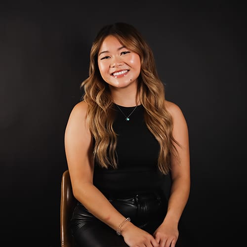 A smiling woman with long, light brown hair wearing a black sleeveless top and seated on a leather chair against a dark background. she has a necklace and a bracelet on.