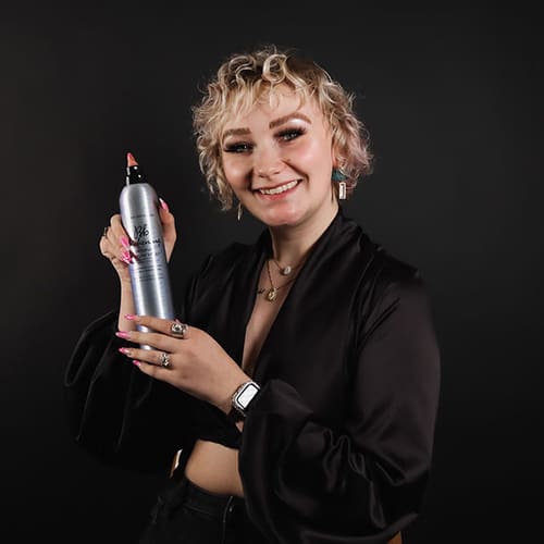 A smiling woman with curly blonde hair, holding a hair product bottle, dressed in a black blouse against a dark background. she wears a watch and has pink nail polish.
