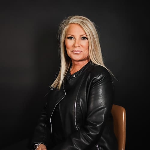 A woman with long blonde hair wearing a black leather jacket, seated against a dark background, looking at the camera with a slight smile.