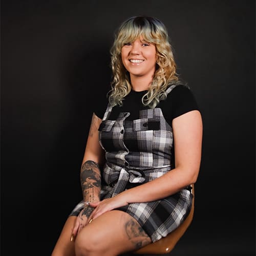 A woman with blonde hair highlighting streaks of blue sits smiling on a chair, wearing a plaid dress over a black shirt, adorned with visible tattoos on her arms.